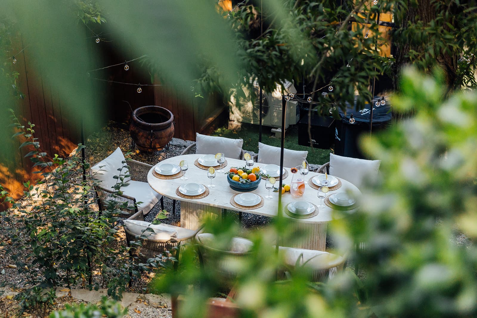 The outdoor dining table set for a group, seen through the garden greenery, string lights above and a bowl of citrus on the table