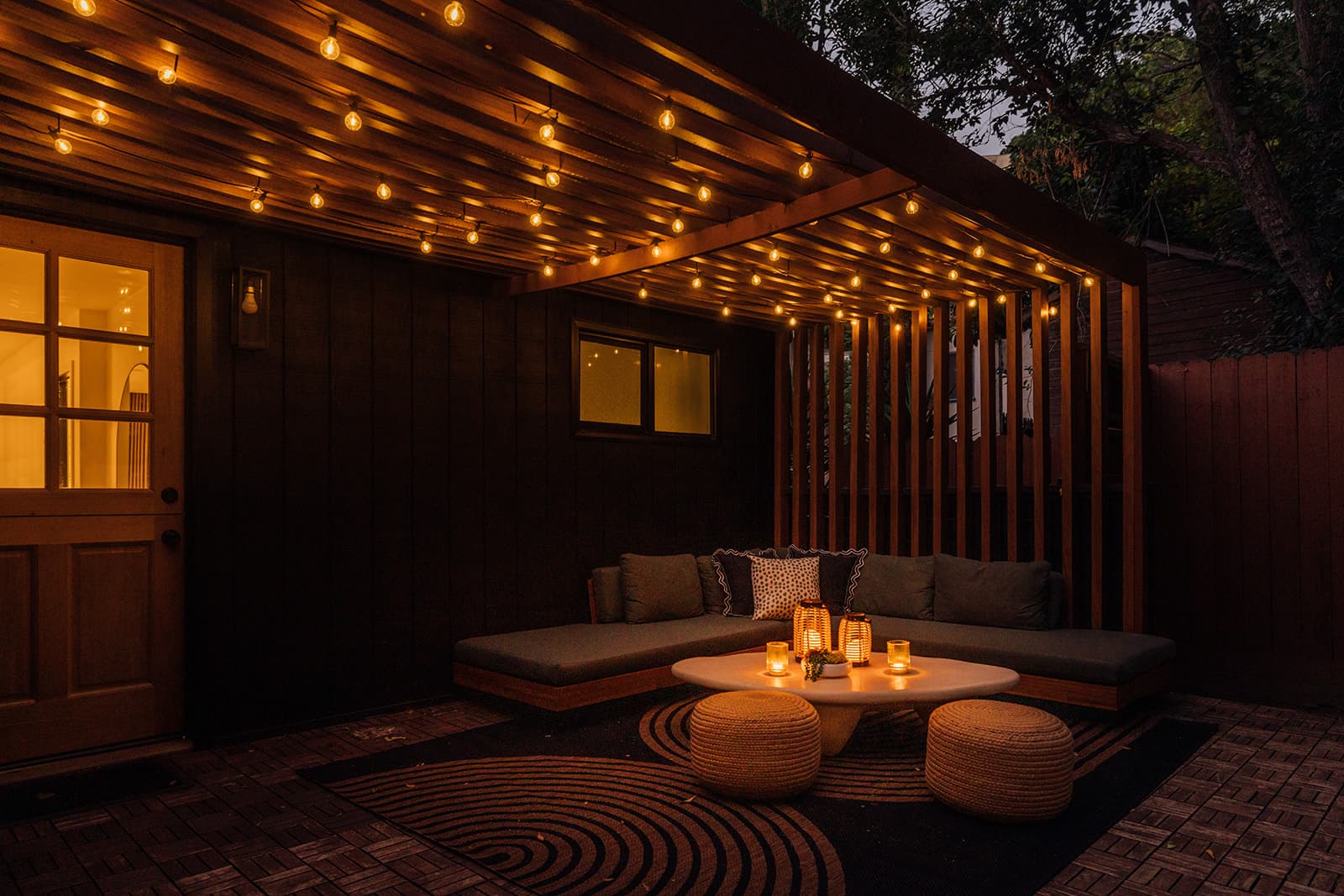 A pergola lounge glowing at night: a green-cushioned sectional and two woven poufs around a low table of lit lanterns, under a wood-slat pergola strung with warm bulbs, beside the house's lit glass door and window
