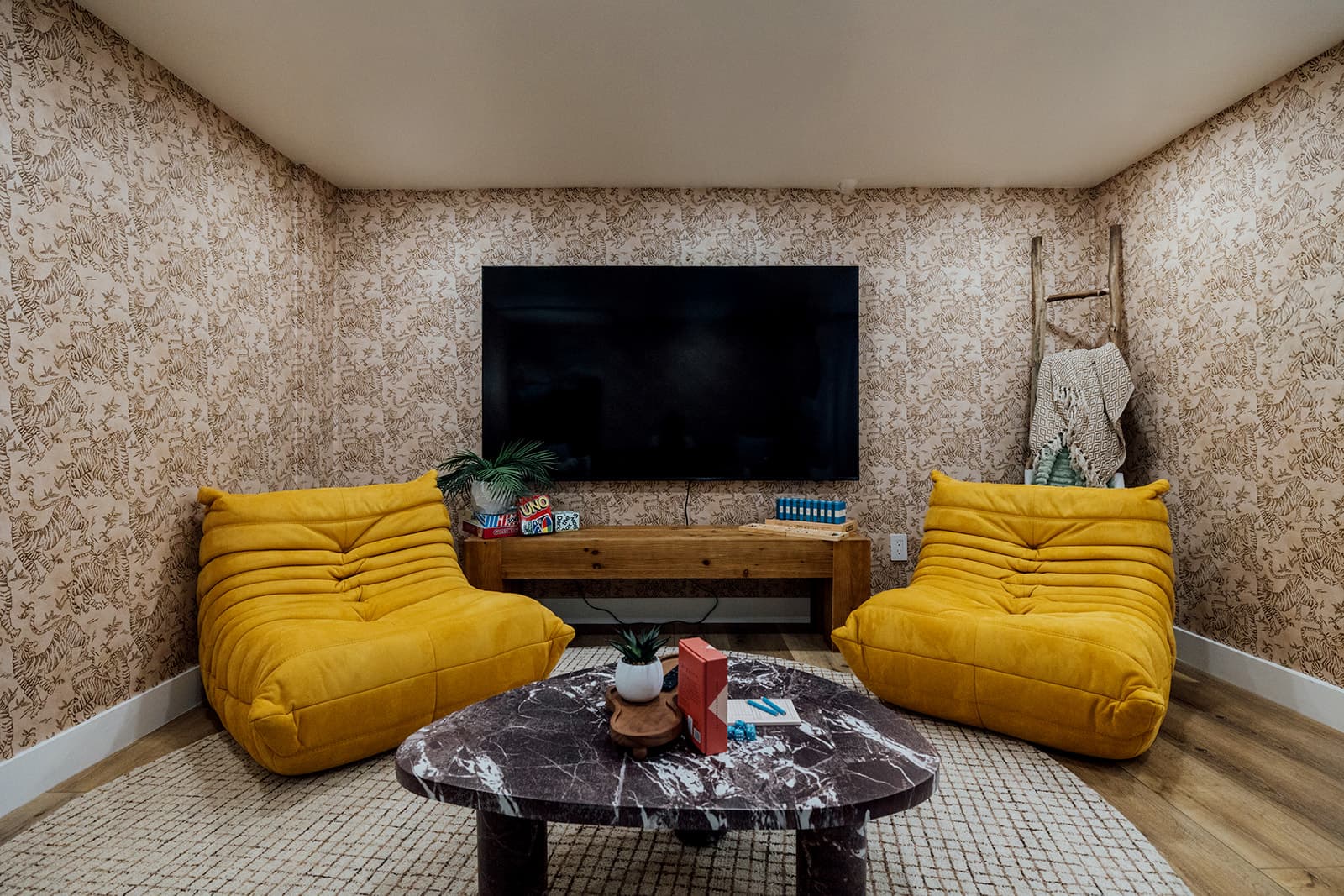 The movie & games lounge: two mustard lounge chairs around a marble table set with board games, a wall-mounted TV, and a wooden blanket ladder