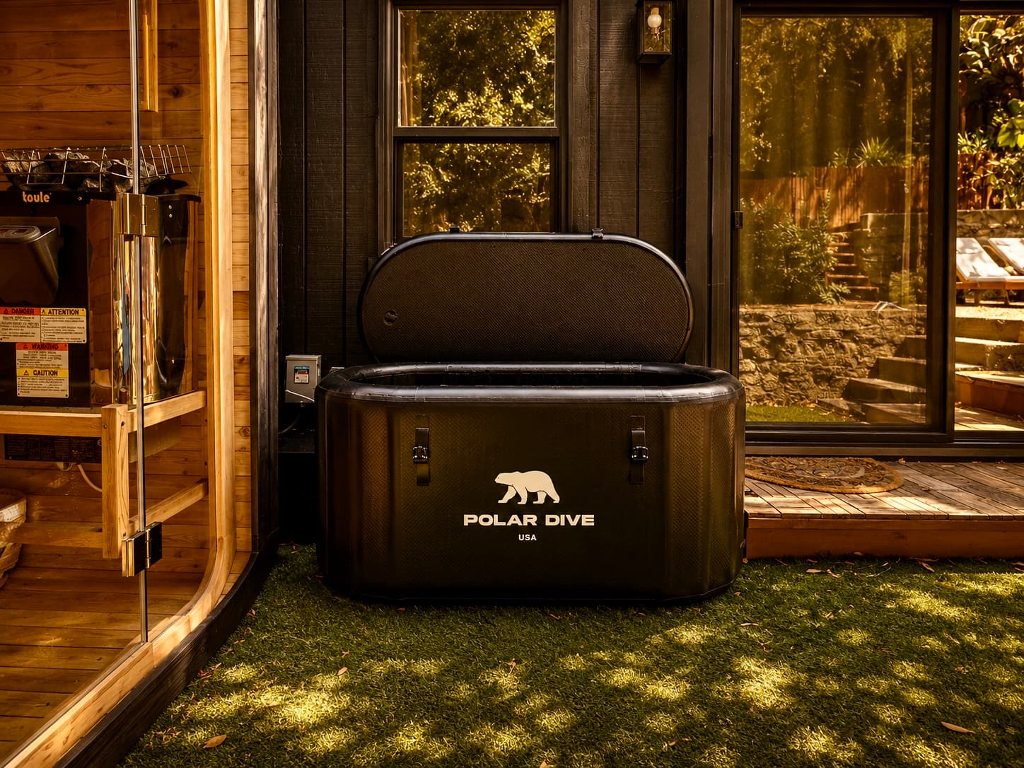 The cold plunge, a black tub on the lawn beside the cedar sauna's glass door and the black board-and-batten house, garden beyond