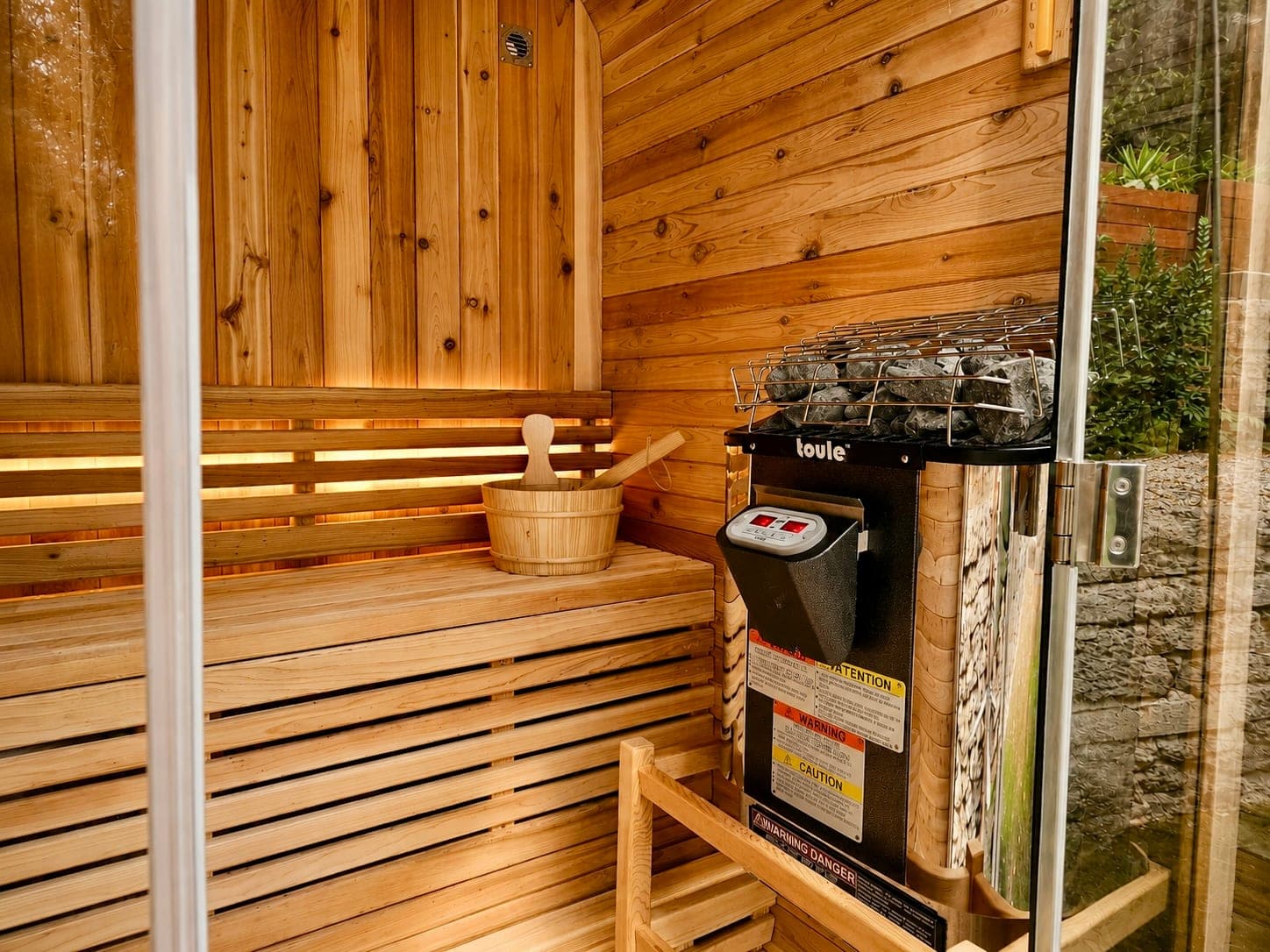 Inside the cedar sauna: slatted cedar benches, a wooden bucket and ladle, and the heater topped with stones