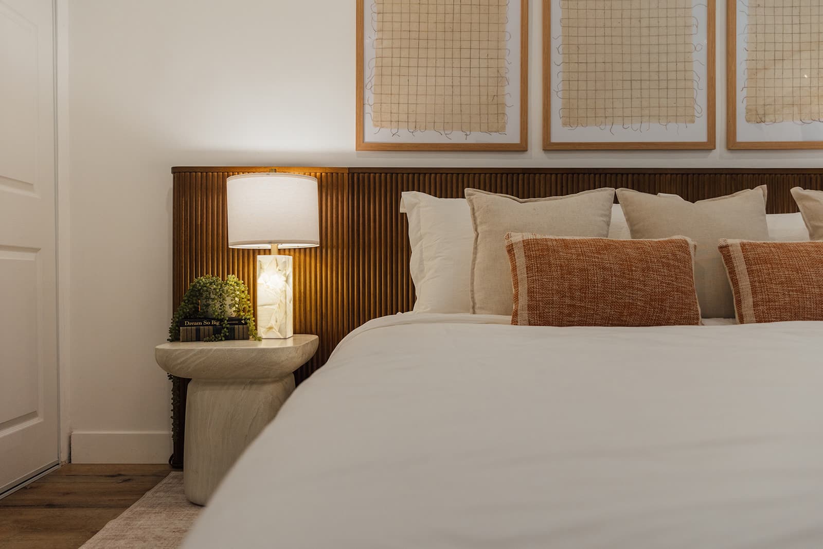 A fluted wood headboard with white and rust textured pillows, a stone table lamp on a curved plaster nightstand