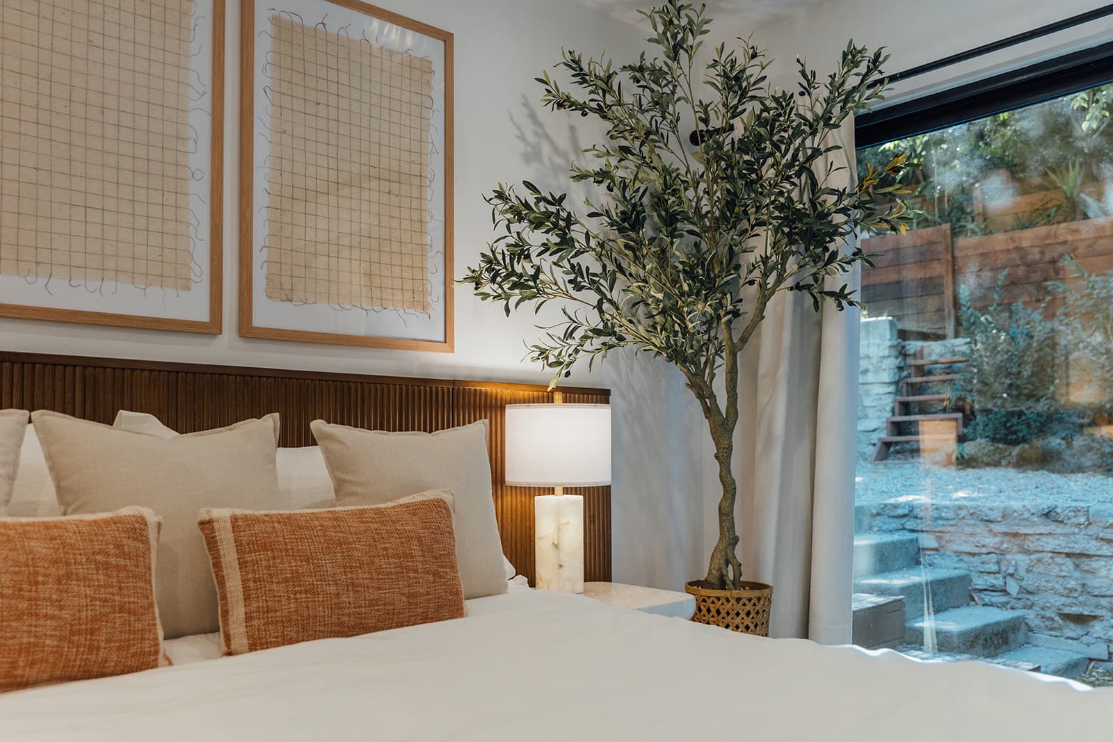 The garden bedroom bed with cream and rust textured pillows, an alabaster lamp and a tall potted olive tree at a floor-to-ceiling window over a stone garden stairway