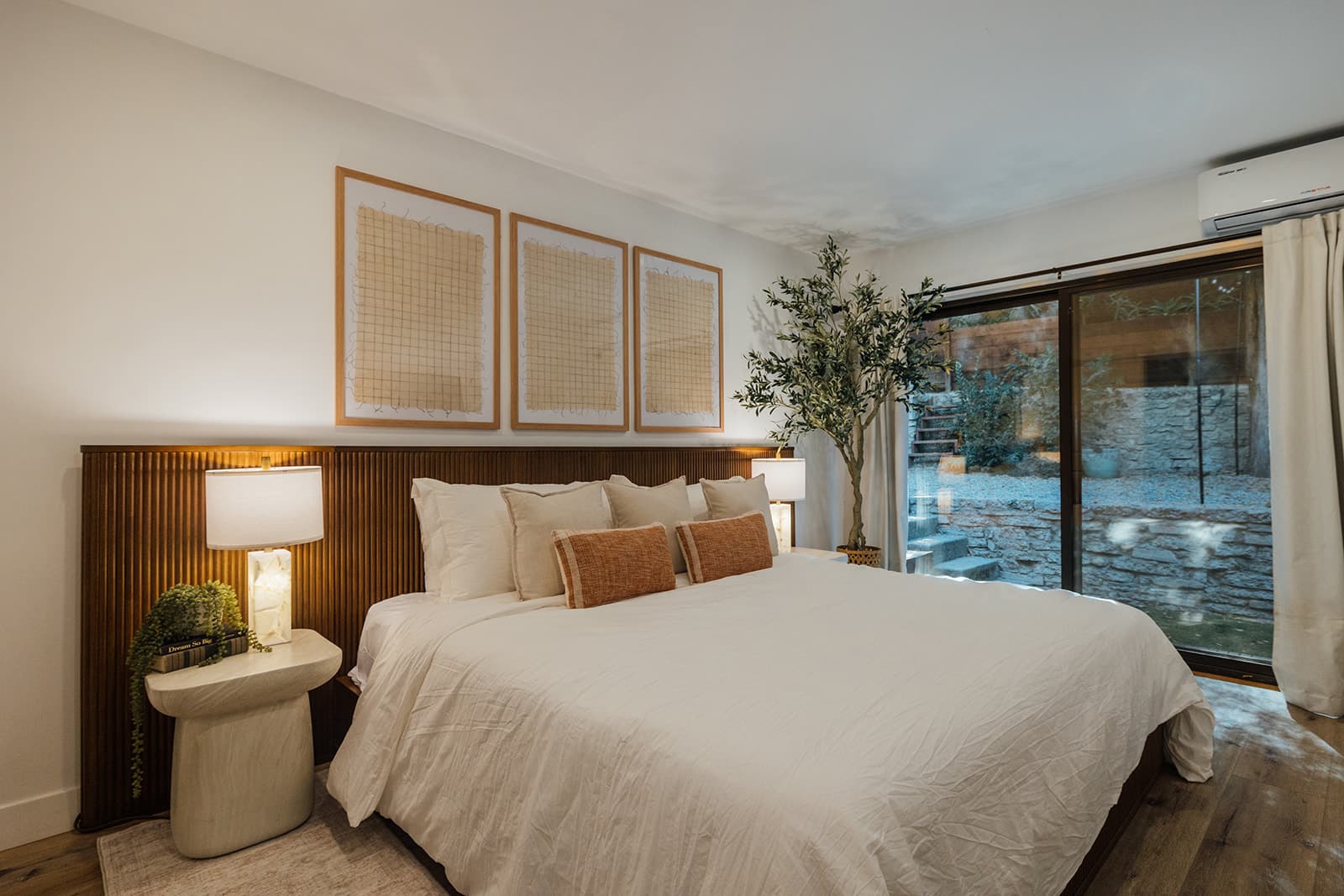 A king bed with fluted headboard and rust lumbar pillows beside a sliding glass door onto a stone-walled garden with steps