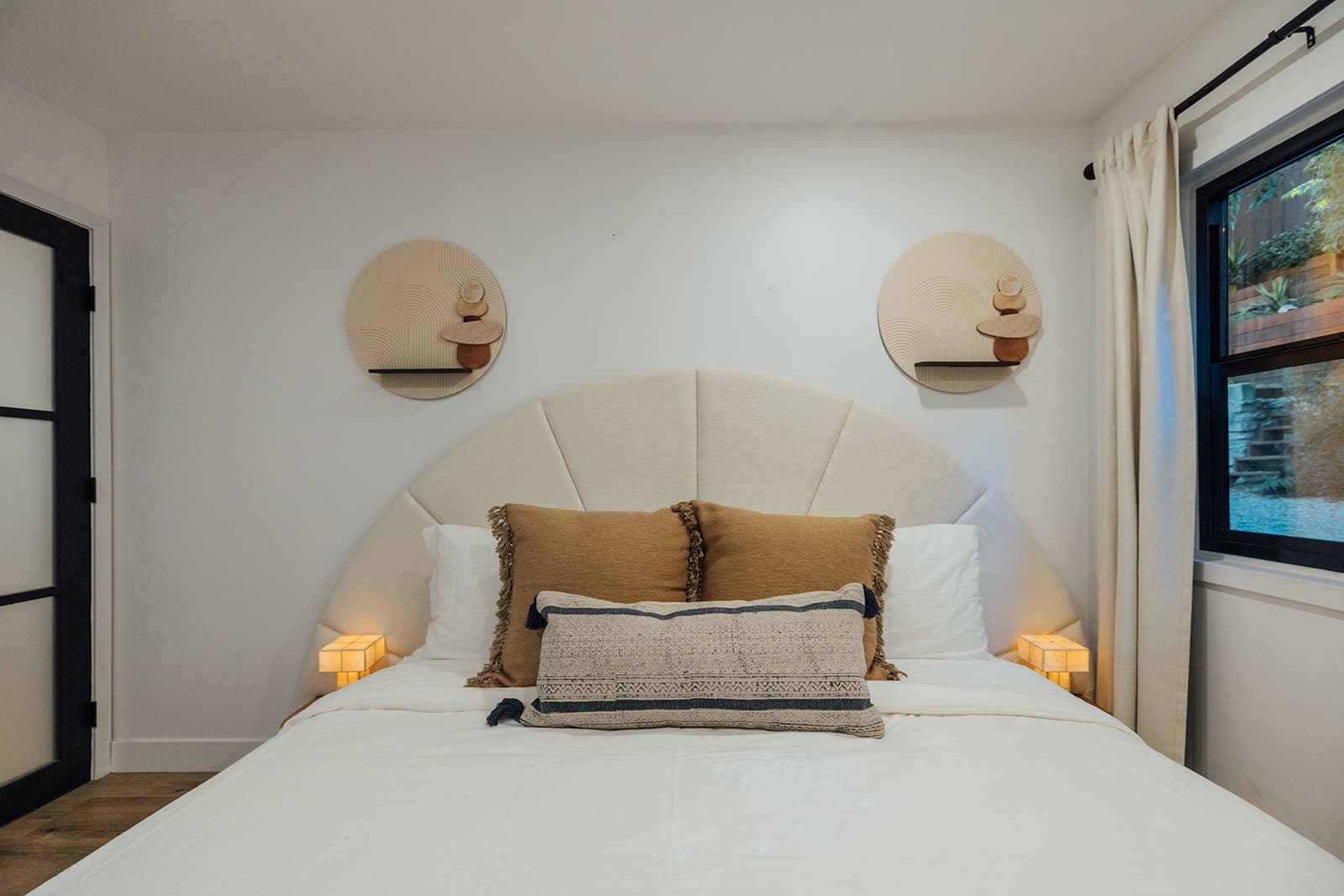 A white-dressed bed with an arched boucle headboard, tan fringed pillows and a navy-striped lumbar, flanked by round wall art and cube lamps