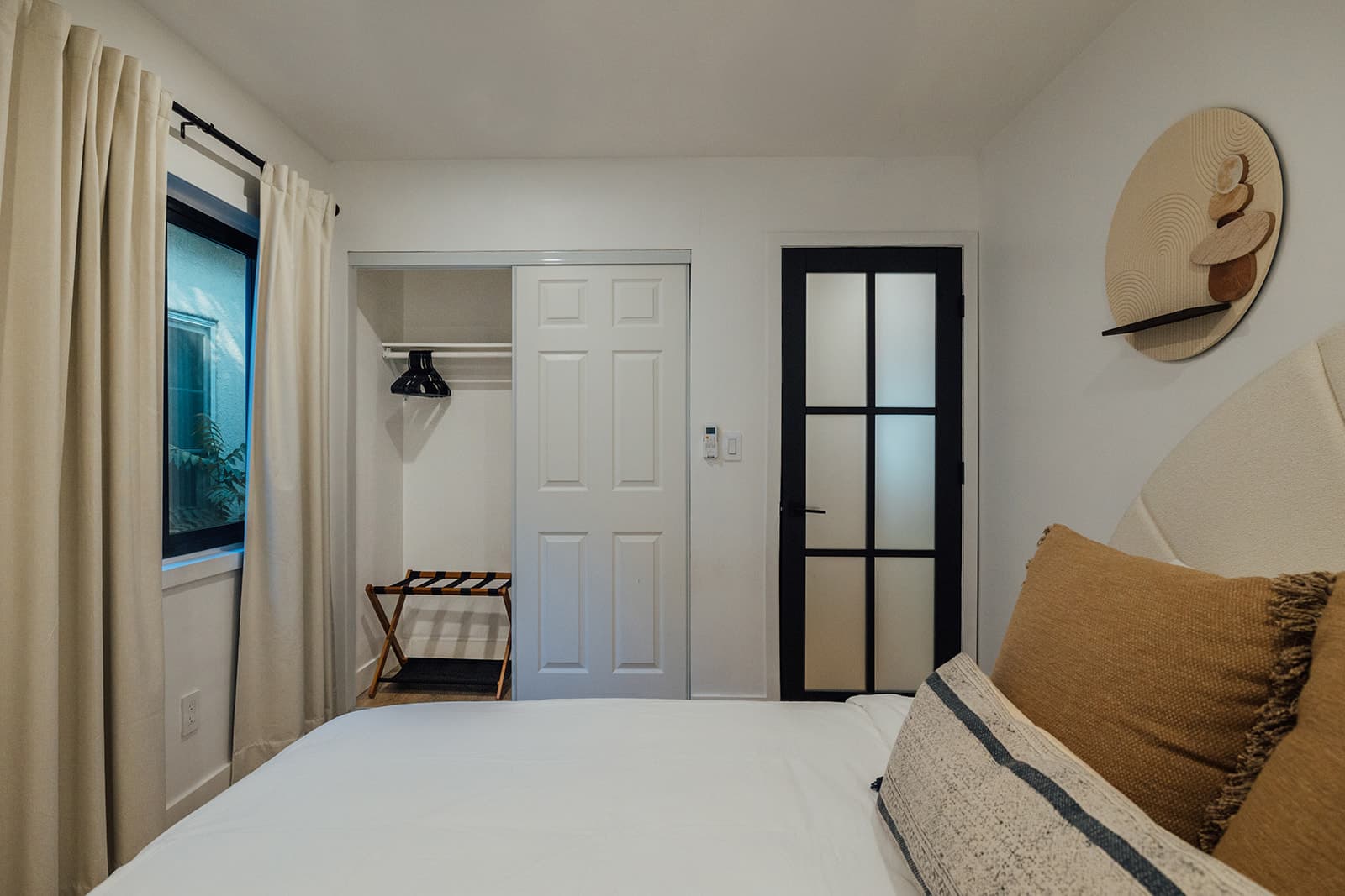The cozy king bedroom with a curtained window, an open sliding-door closet with a luggage rack, a black-framed glass door and a round textured wall disc above the upholstered headboard
