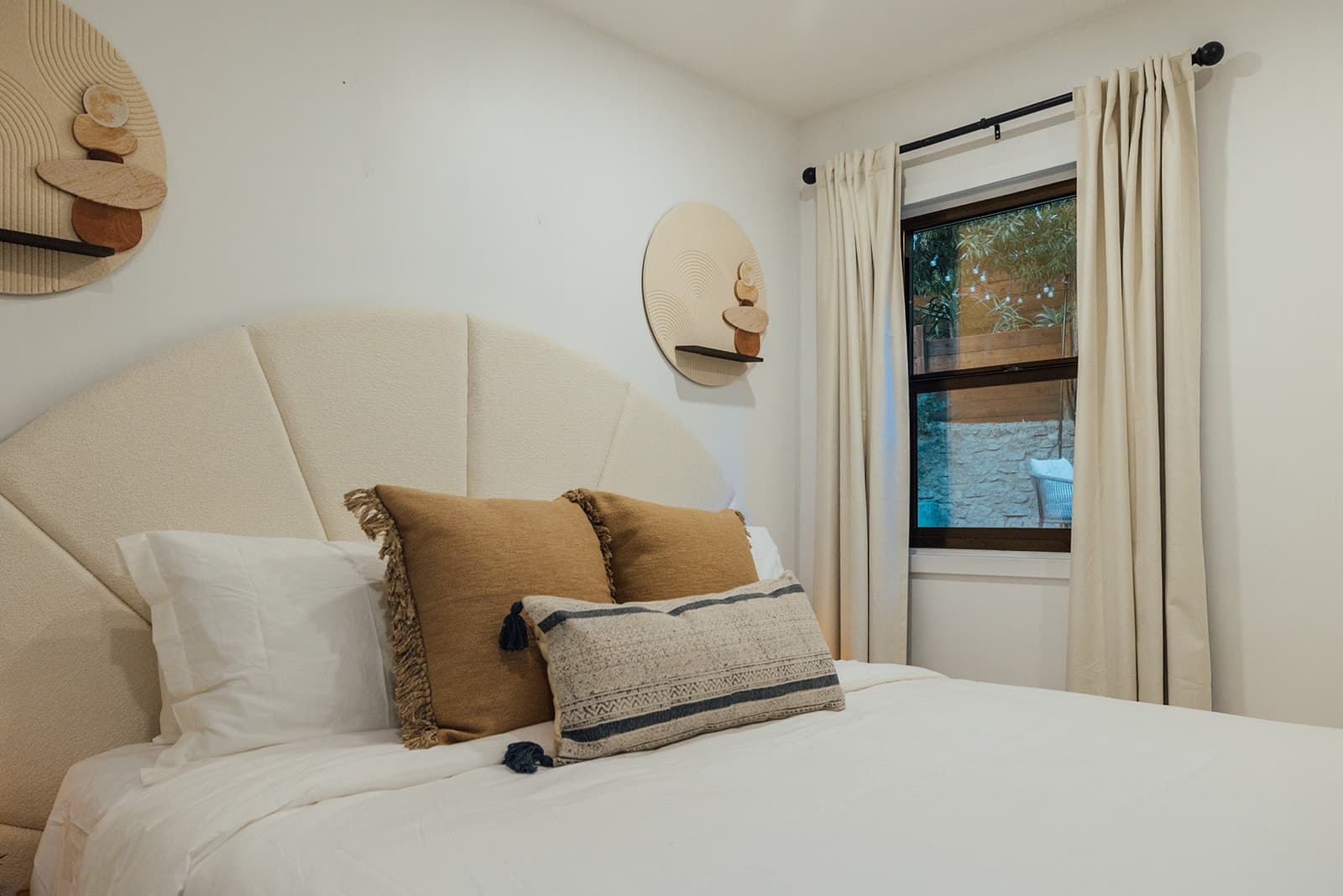 The bed with arched boucle headboard and tan fringed pillows beside a black-framed window with cream curtains and a garden view