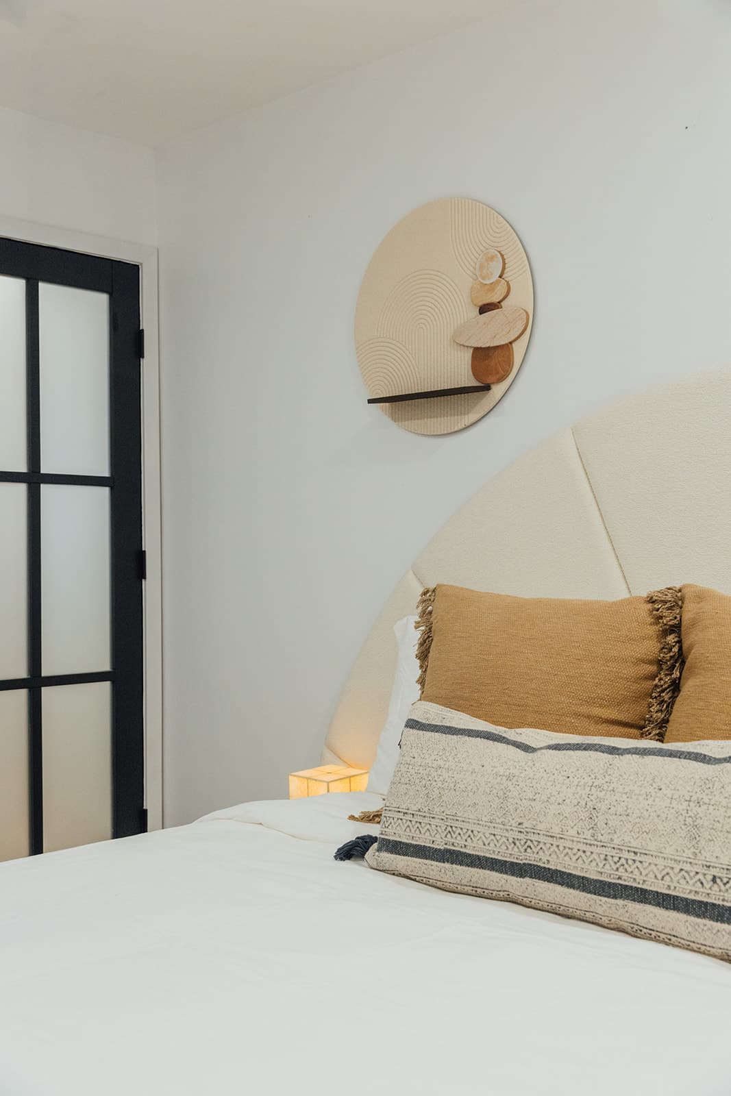 The arched boucle headboard with tan and striped pillows, a circular wall shelf of wood objects and a black-framed glass door alongside