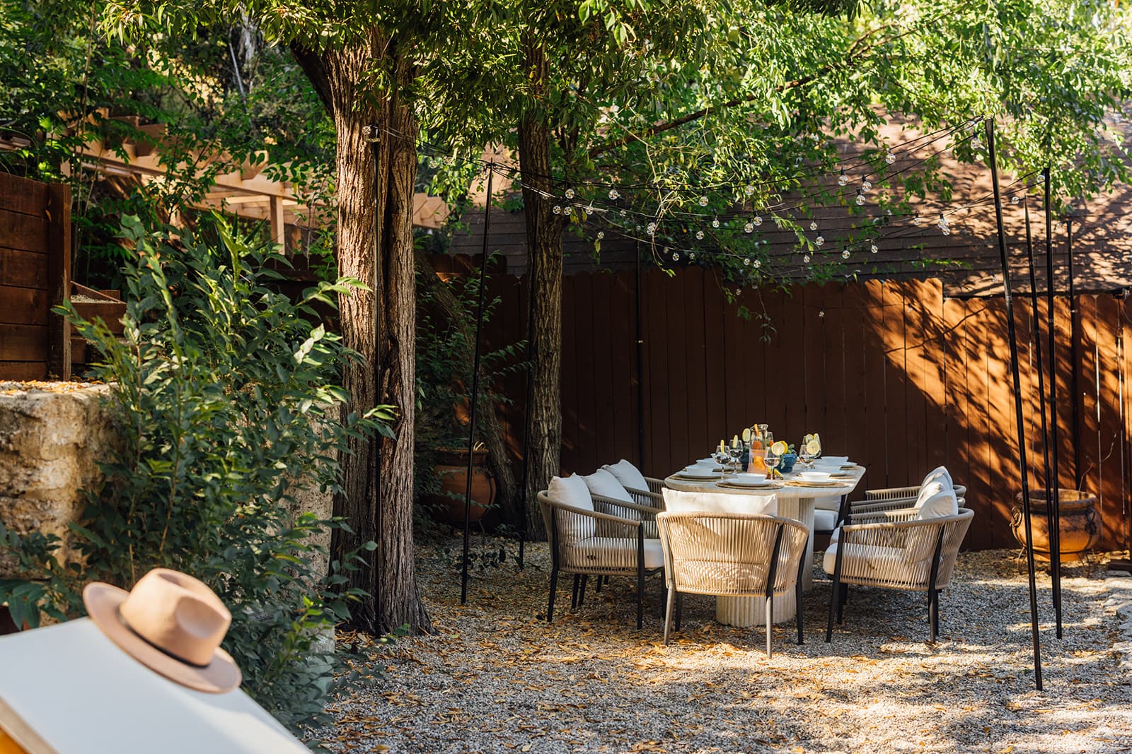 A round travertine dining table set with plates and glasses, ringed by woven rope armchairs beneath tall trees and string lights, a straw hat on a lounger in front