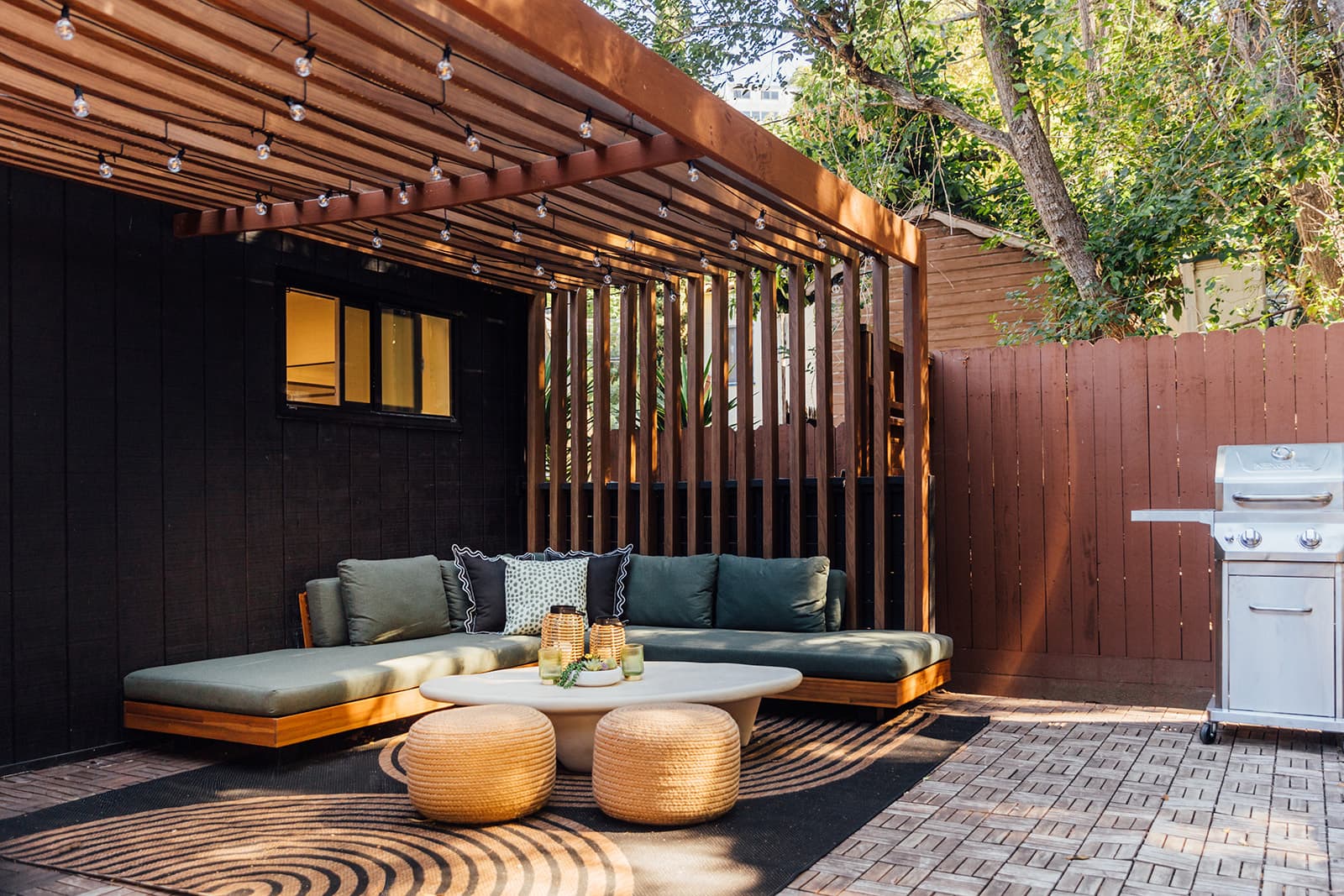 A covered lounge under a wood-slat pergola strung with cafe lights, a green-cushioned corner sofa, two rattan poufs on a patterned rug and a gas grill to the right