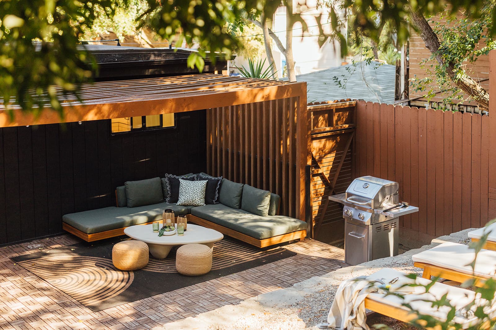 An elevated view of the green sectional lounge with poufs and a coffee table beside a stainless grill and wooden loungers, framed by trees and a slatted pergola
