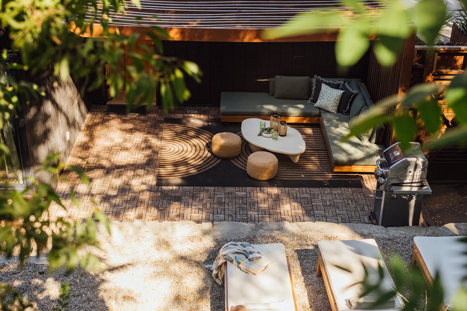 An overhead view through leaves of the green sectional, round coffee table and woven poufs on a patterned rug, with a grill and cushioned loungers on the gravel below