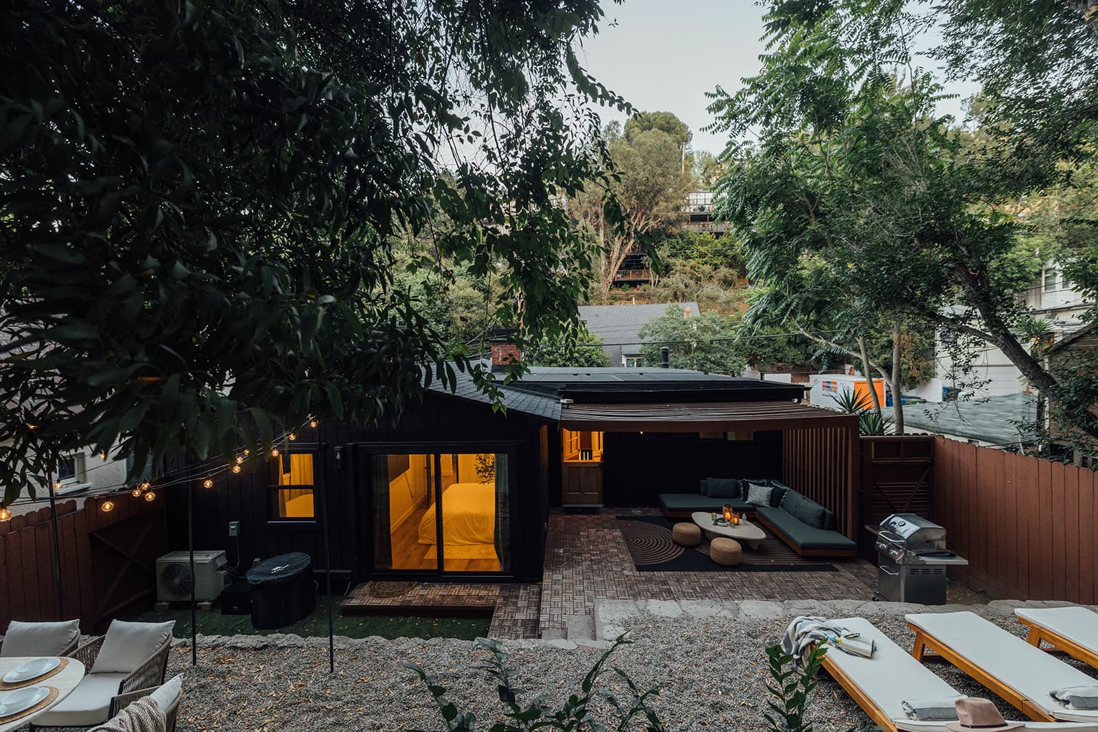An elevated view of the garden at dusk: the black board-and-batten house with a warmly lit bedroom, the pergola lounge, dining set and loungers, string lights and hillside trees