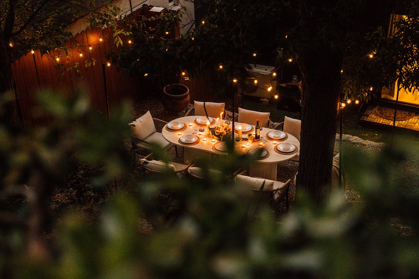 An oval stone table set with white plates, lit candles, a bowl of lemons and a wine bottle, ringed by rope chairs beneath string lights strung between trees at dusk