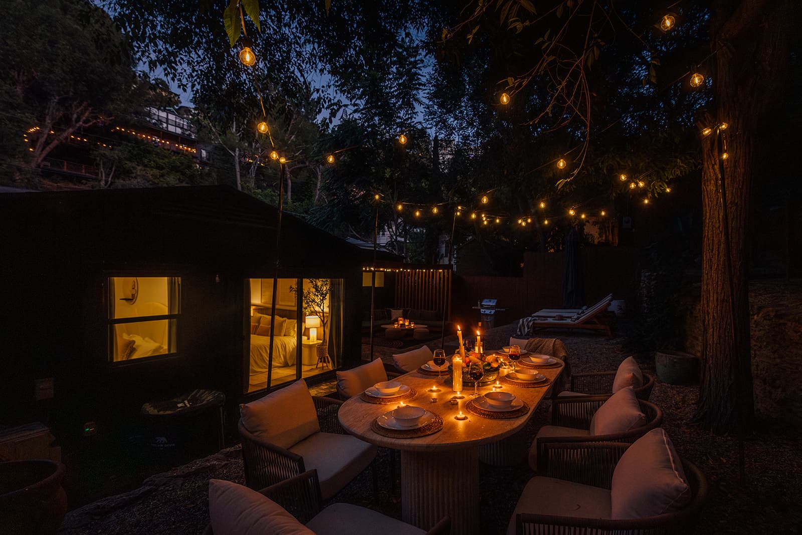 A nighttime backyard with cafe lights overhead illuminating a candlelit dining table and chairs, a lit bedroom glowing through glass doors, loungers and a grill in the distance
