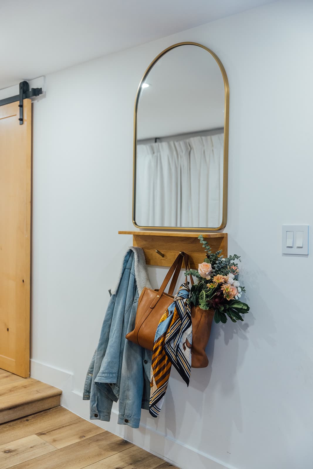A brass arched mirror on a wood shelf beside a barn door, with a denim jacket, a tan leather tote, a printed scarf and a flower bouquet hanging below