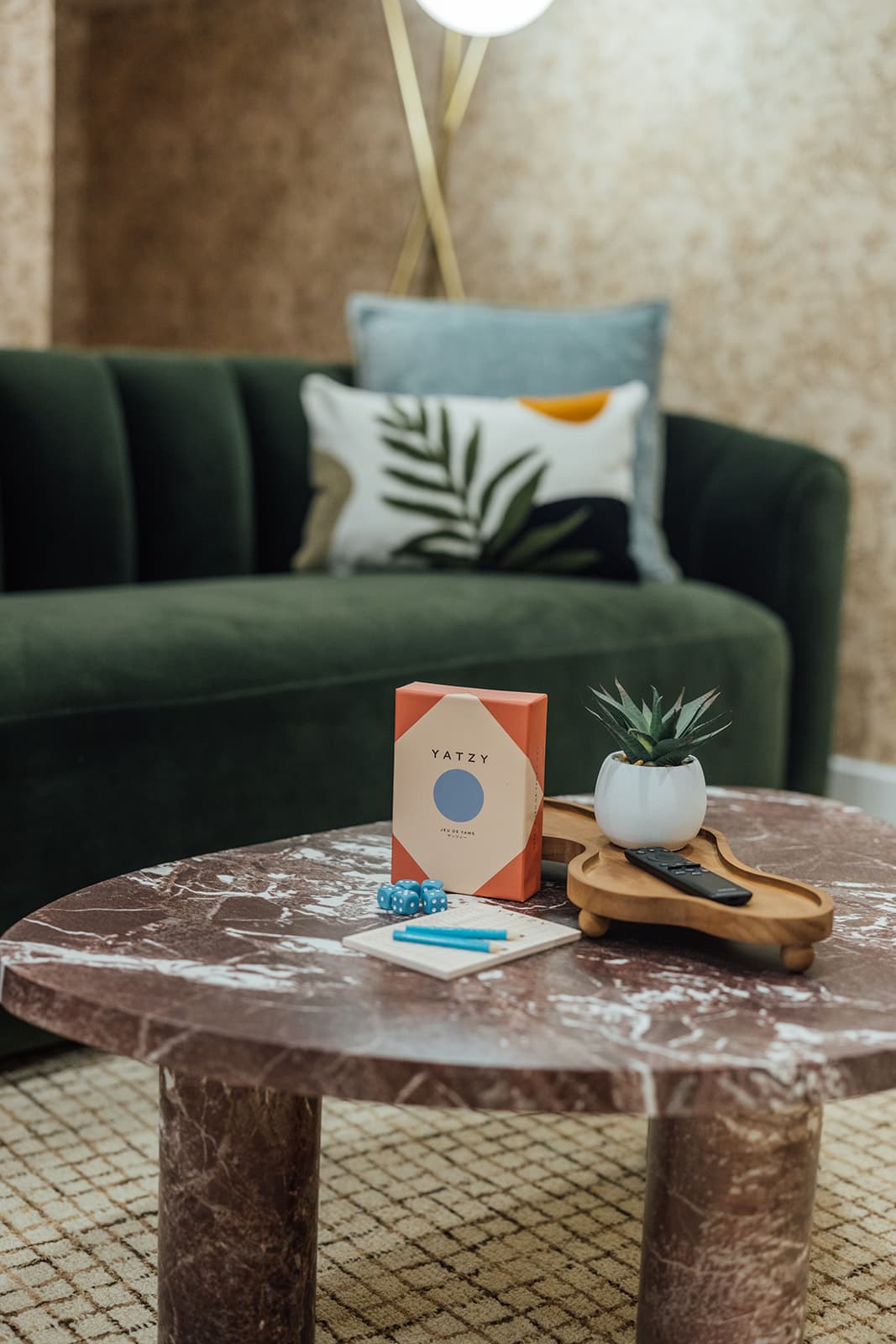 A dice game with blue dice and a scorepad on a round burgundy marble table, a potted succulent on a tray and the dark green velvet sofa behind