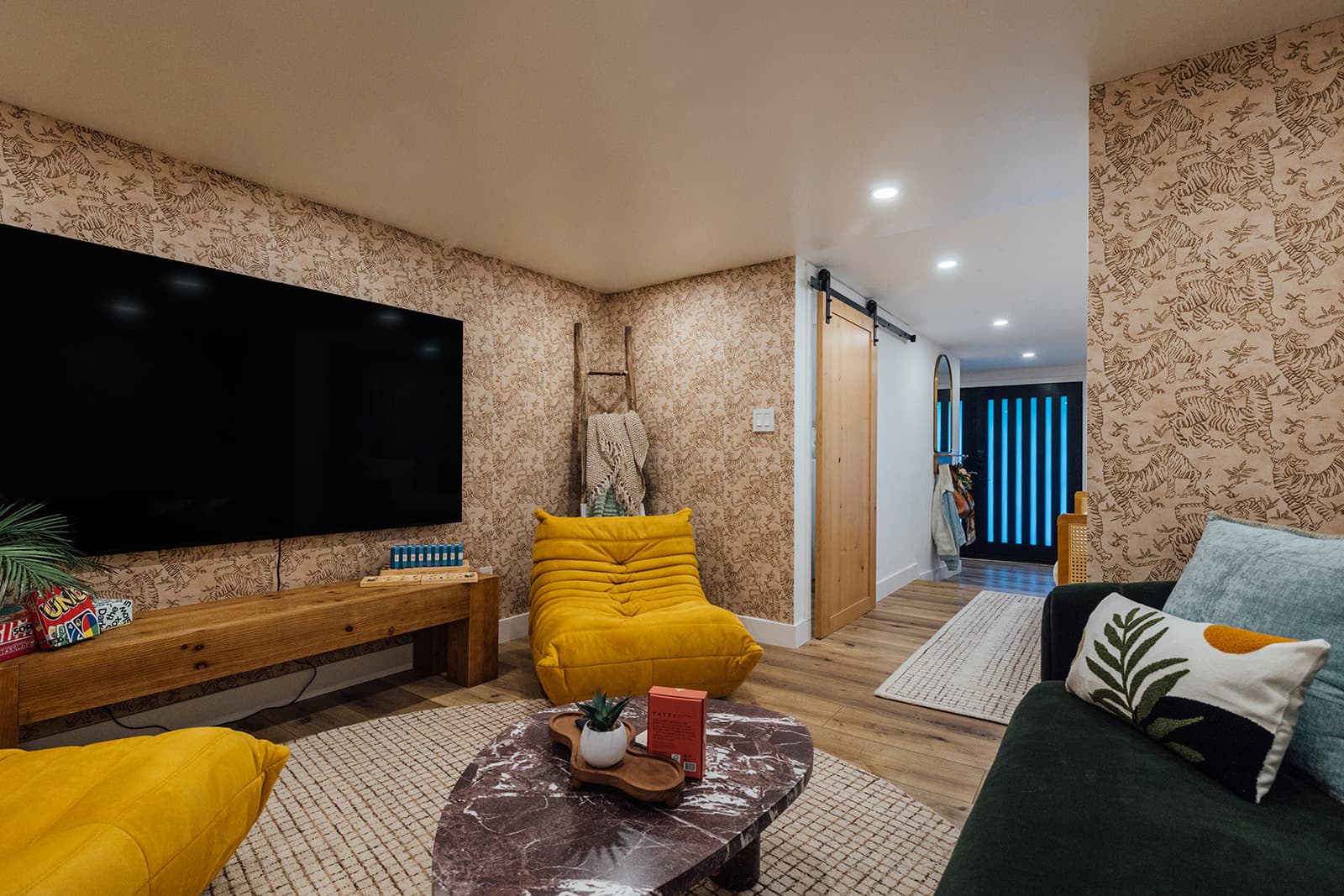 A wide lounge view with a wall-mounted television over a console of games, a mustard Togo chair, a burgundy marble table and a green velvet sofa toward a blue-lit entry