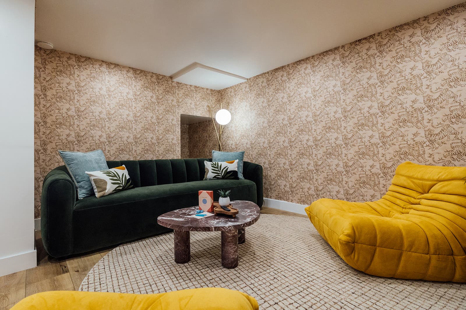 A dark green velvet sofa with palm-print cushions against tiger-print wallpaper, facing a burgundy marble coffee table and a mustard Togo floor sofa