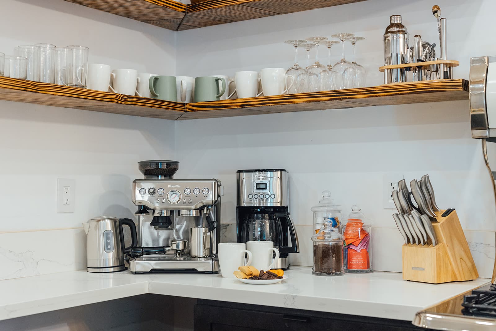 A stainless espresso machine, drip coffee maker, and kettle on the white quartz counter, mugs and glasses on the wood shelves above