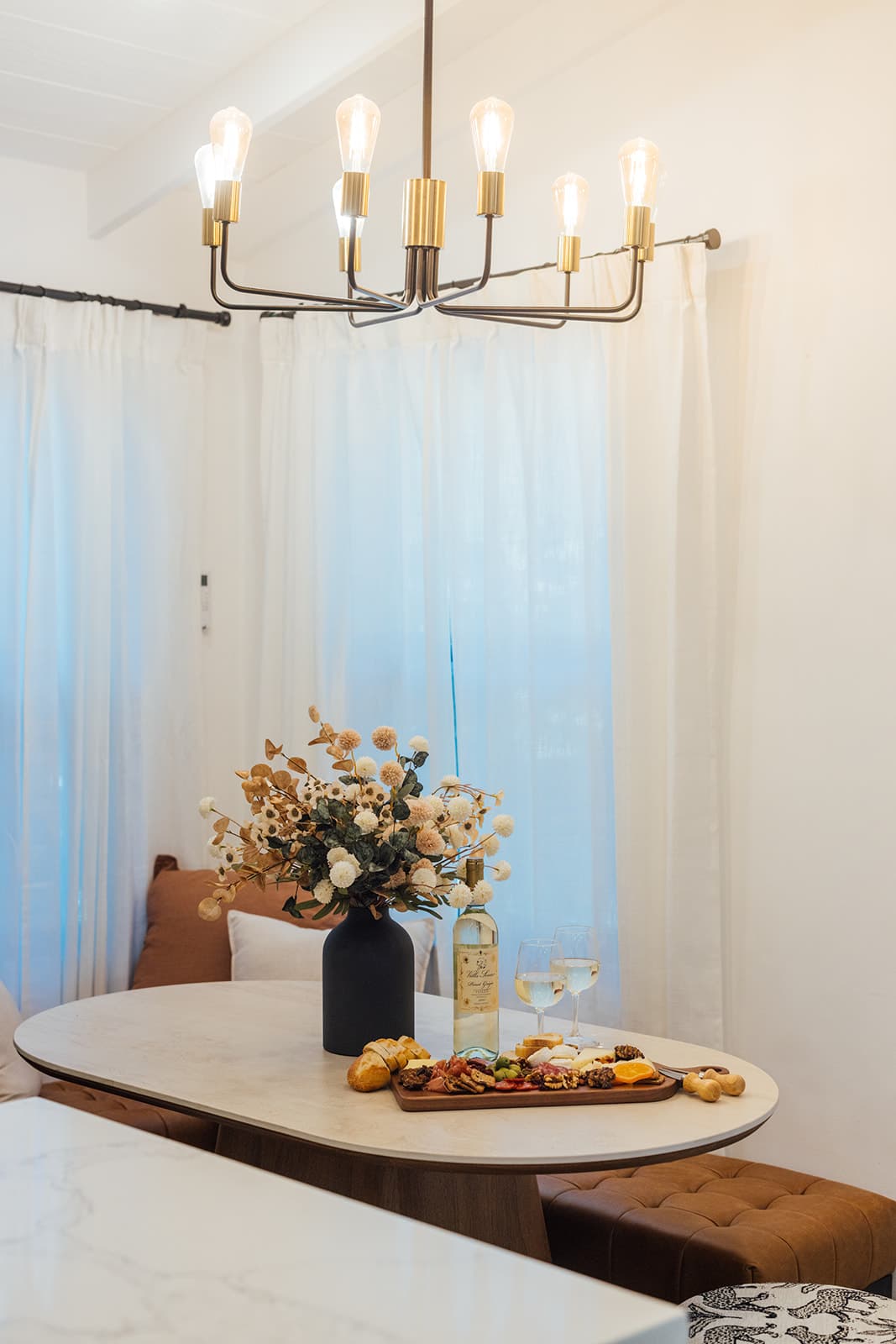 An oval stone-top table set with a charcuterie board and white wine beneath a brass Edison-bulb chandelier, sheer curtains and a tan leather bench behind