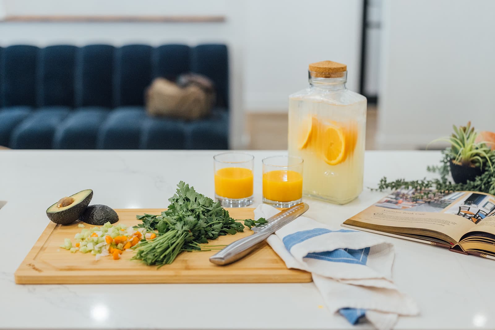 A bamboo board of halved avocado and chopped vegetables beside two glasses of orange juice on the white quartz island, the navy tufted sofa behind