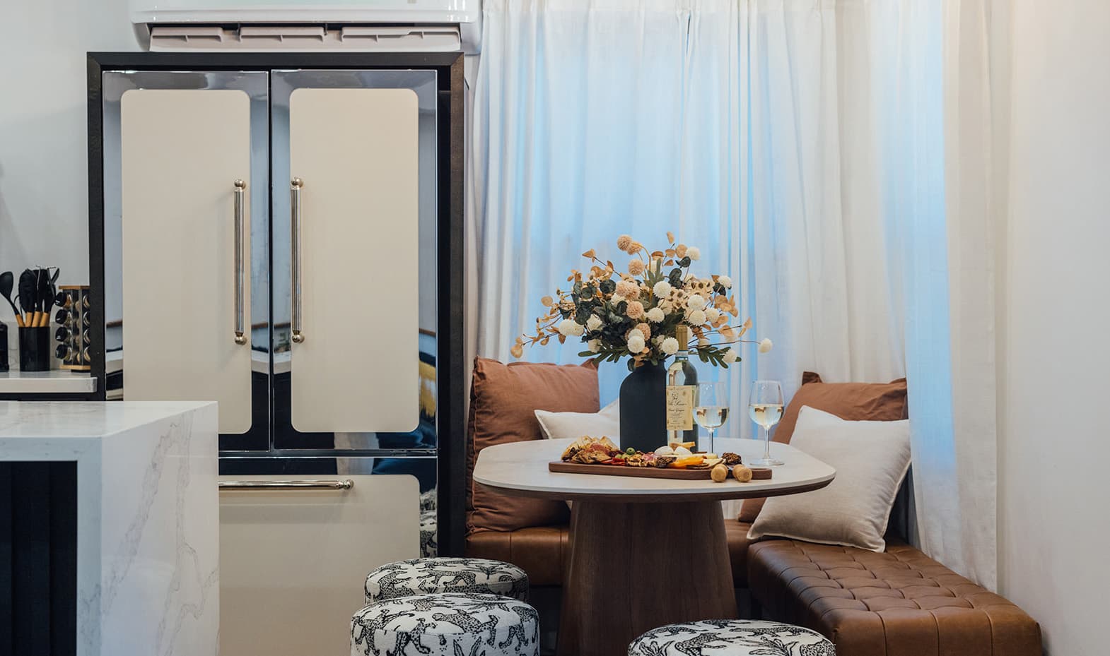 A round wood table with a charcuterie board and dried flowers against a tan leather banquette, beside a cream double-door refrigerator and leopard-print stools