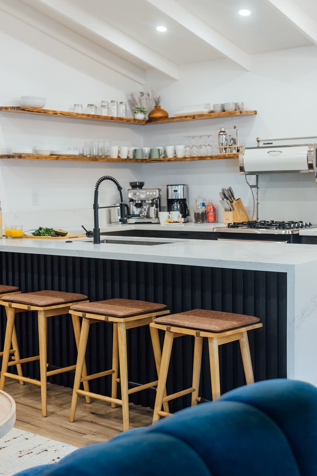 Leather-cushioned wood stools along a black fluted kitchen peninsula, the marble counter and burnt-wood open shelving beyond, a blue velvet sofa in the foreground