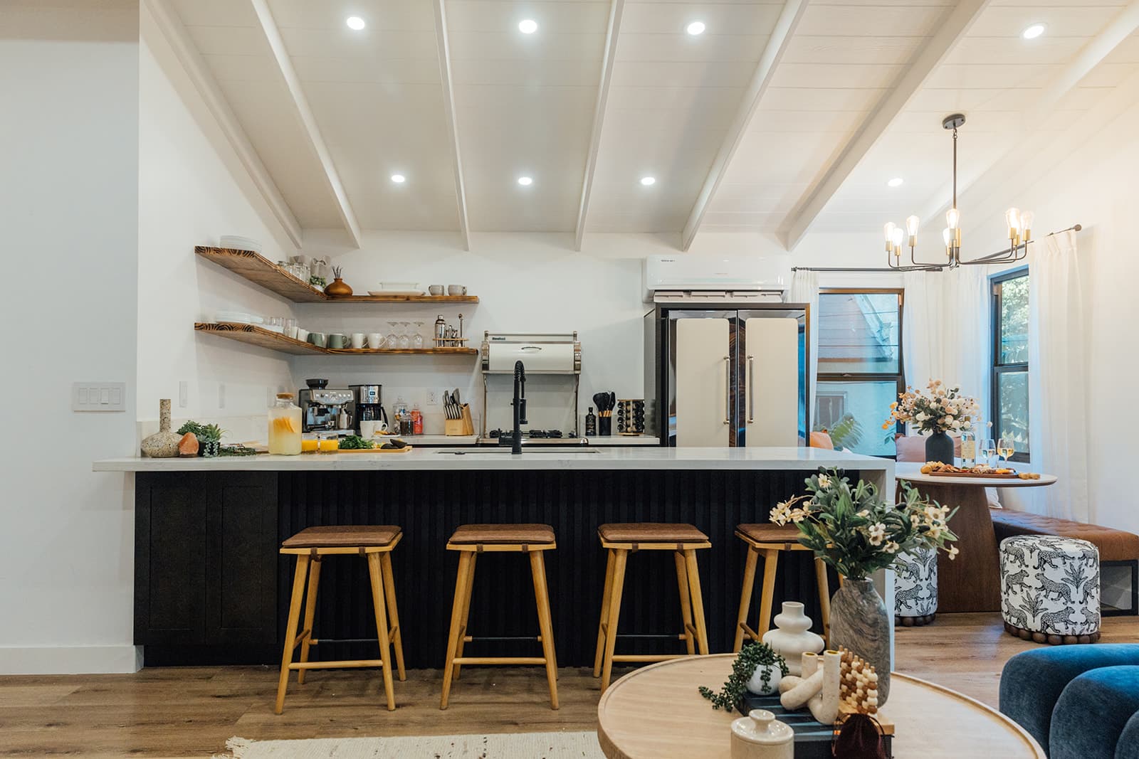 The kitchen with a marble-waterfall island lined with wood stools, open shelving and a dining nook under a brass chandelier below the white beamed ceiling