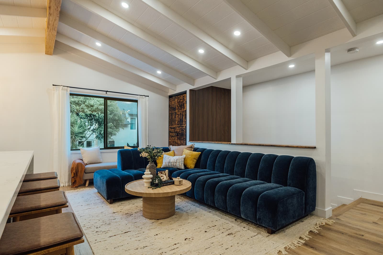 An open living area with a navy velvet sectional, a round wood coffee table and a cream fringed rug under a white beamed ceiling, beside the kitchen peninsula stools
