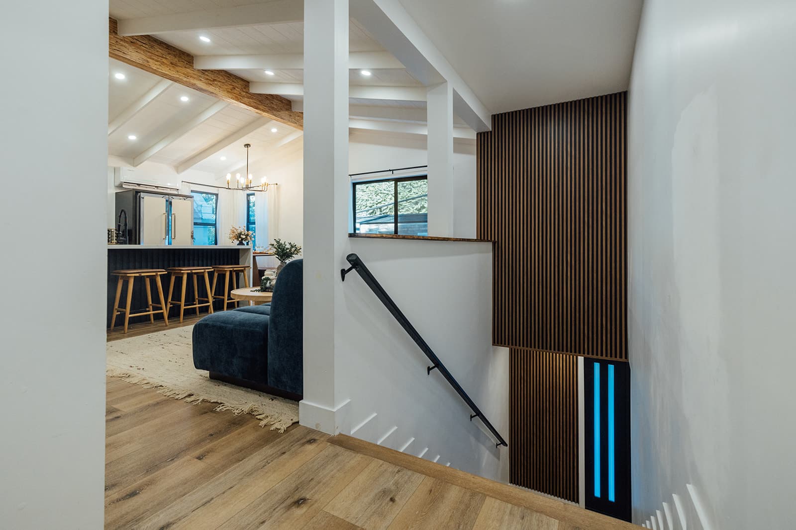 A view down a wood-slat stairwell lit with blue accent lighting toward the kitchen peninsula, bar stools and chandelier beyond