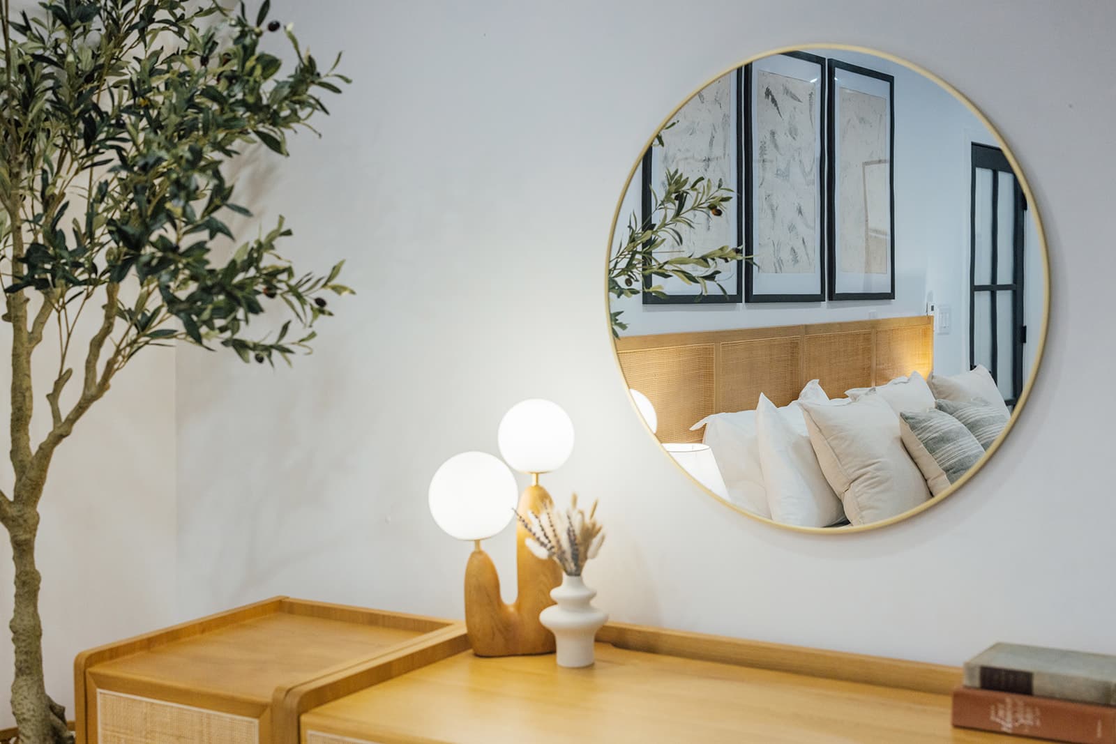 A gold-framed round mirror over a cane console holding a double-globe lamp and dried flowers, with a potted olive tree alongside and the bed reflected in the glass