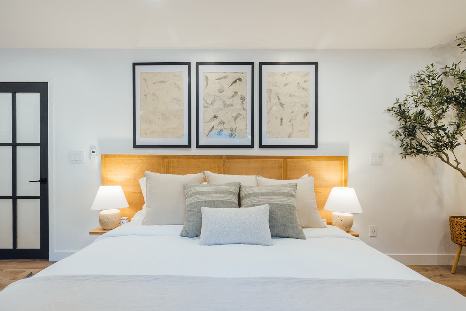 A king cane bed below three framed botanical prints, flanked by matching speckled-ceramic table lamps and a black glass-panel door at left