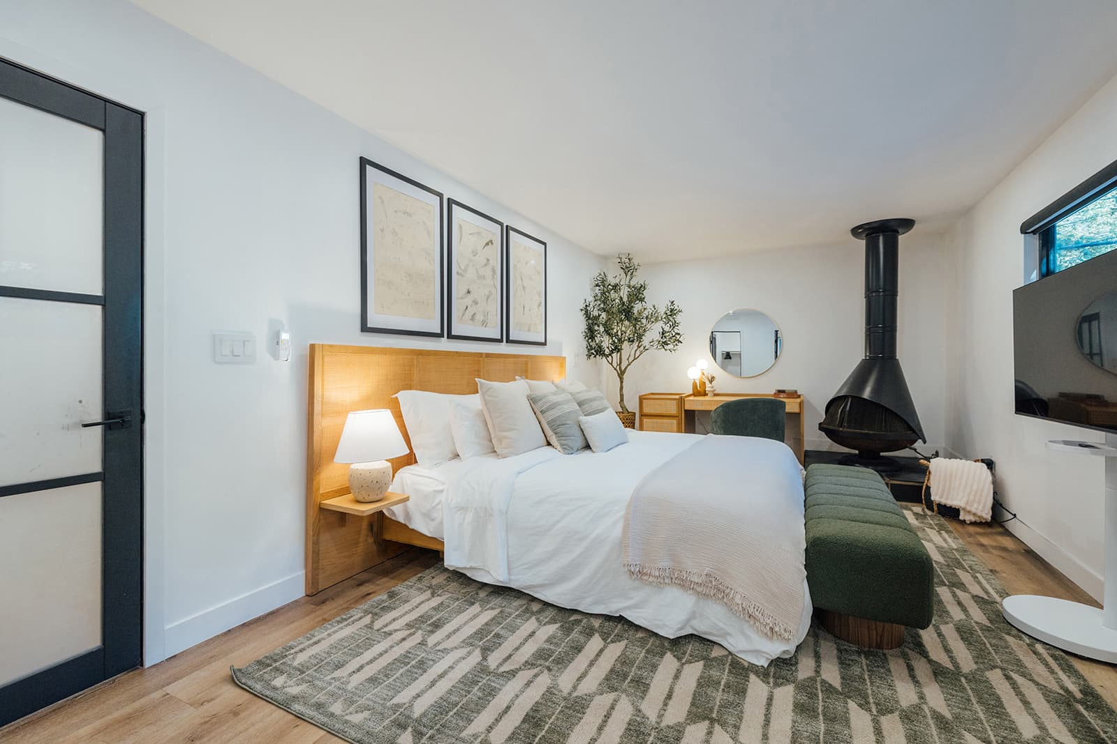 A white-dressed king bed with a cane headboard and framed prints, a green boucle bench at the foot and a black cone fireplace in the corner