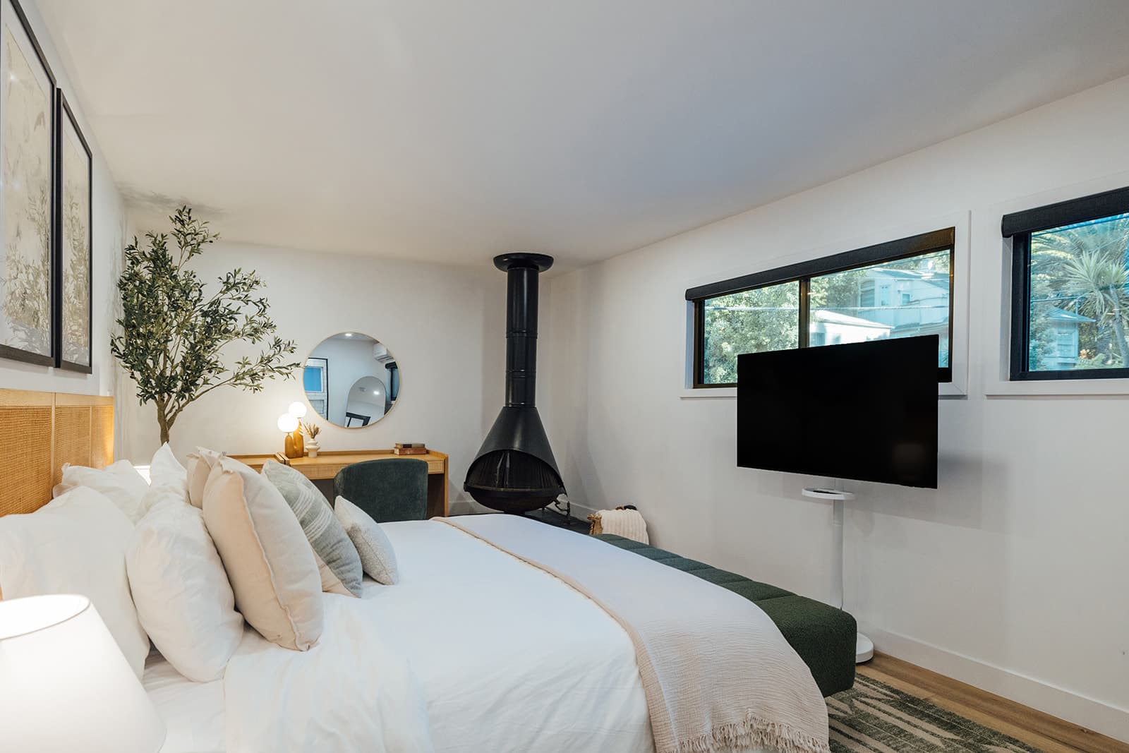 The primary bedroom across the bed toward a wall-mounted television, the black cone fireplace and two windows onto greenery