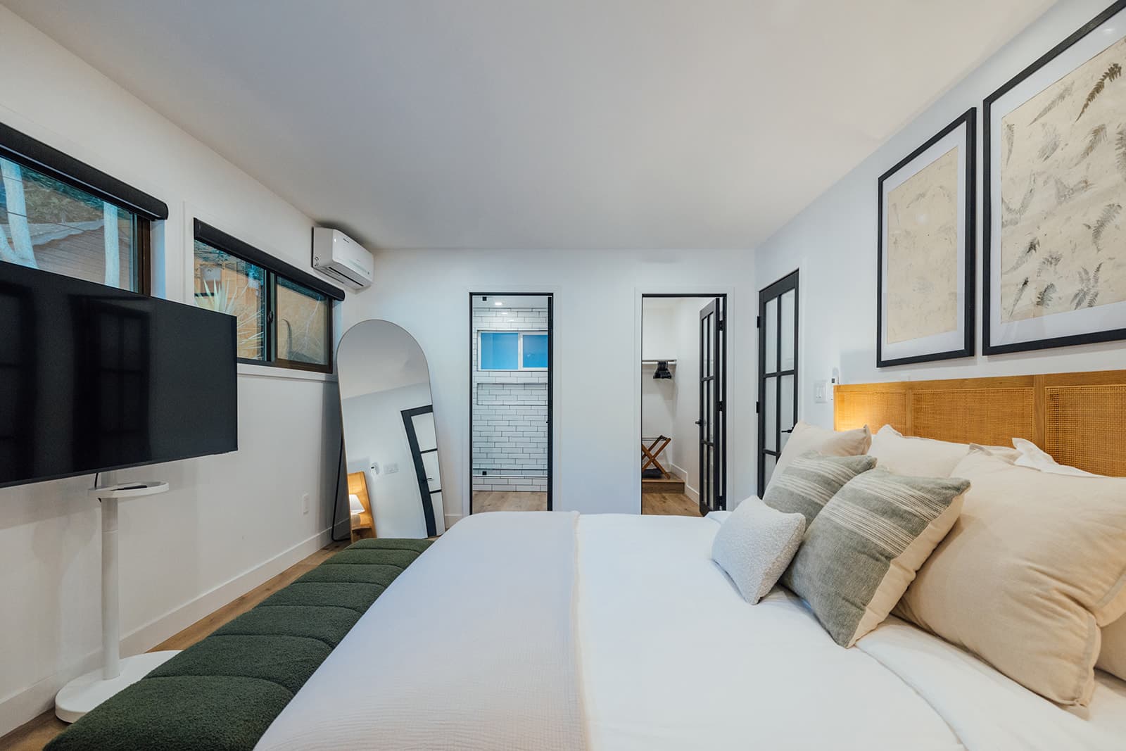 Looking down the bed toward a tiled ensuite, a walk-in closet, an arched floor mirror and a wall-mounted television, framed prints above the cane headboard at right