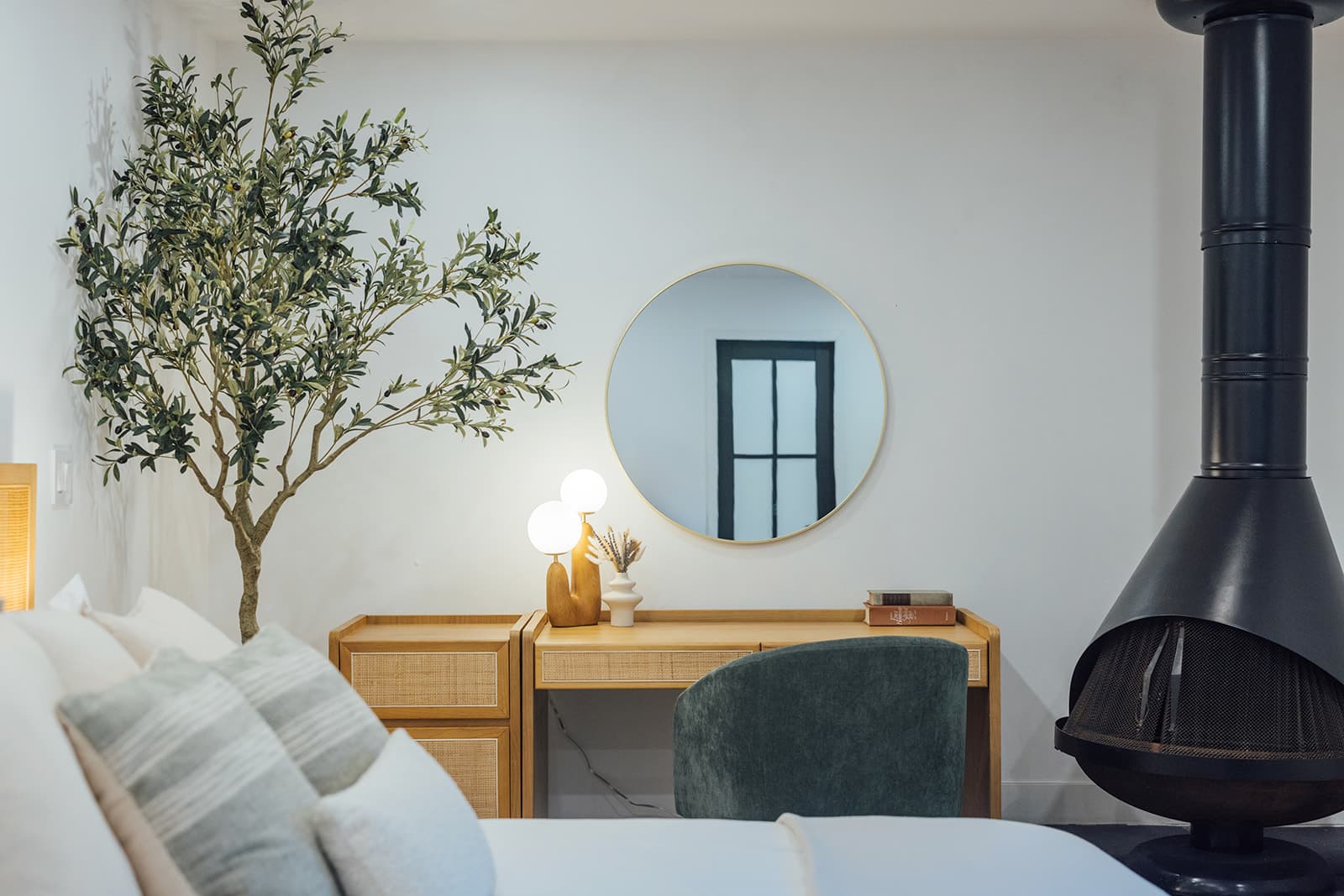 The cane vanity with a green velvet chair, round gilt mirror and globe lamp beside the black cone fireplace and the olive tree